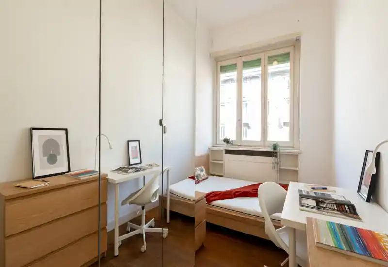 Camera da letto luminosa e compatta con letto singolo, scrivania e armadio con ante a specchio; arredi dal design pulito e luce naturale da una grande finestra.