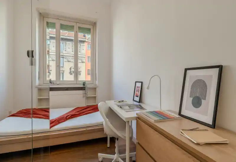 Camera da letto piccola e luminosa con letto singolo ben sistemato, scrivania compatta e luce naturale da una grande finestra. Arredi puliti in stile scandinavo e quadri rendono lo spazio tranquillo e funzionale, ideale per un affittuario singolo.