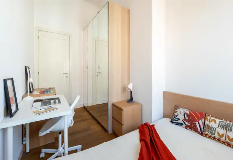 Camera da letto luminosa e compatta con testiera in legno, cuscini decorativi e un plaid rosso; armadio con ante a specchio e scrivania ordinata per lavoro e spazio guardaroba.
