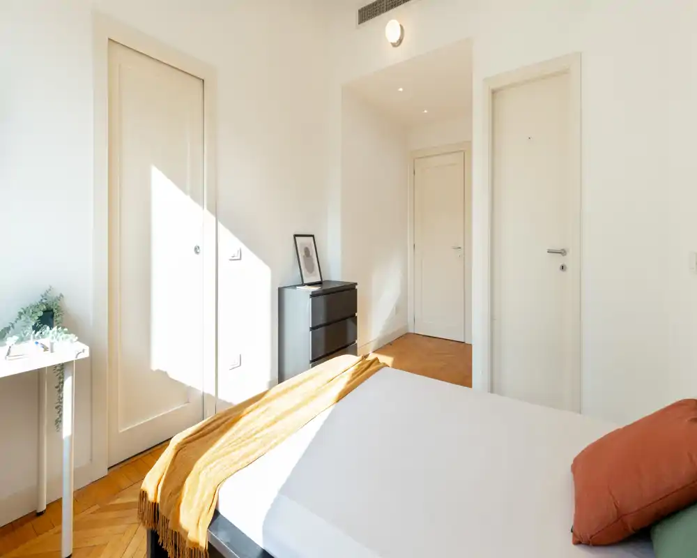 Dormitorio luminoso y minimalista con cama bien hecha, manta cálida y luz natural; paredes blancas y suelo de parquet crean un ambiente moderno y sereno.