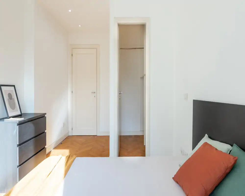 Dormitorio luminoso y minimalista con cabecero visible, cojines de colores y cómoda; paredes neutras y suelo de madera crean un ambiente tranquilo y aireado perfecto para anuncios.