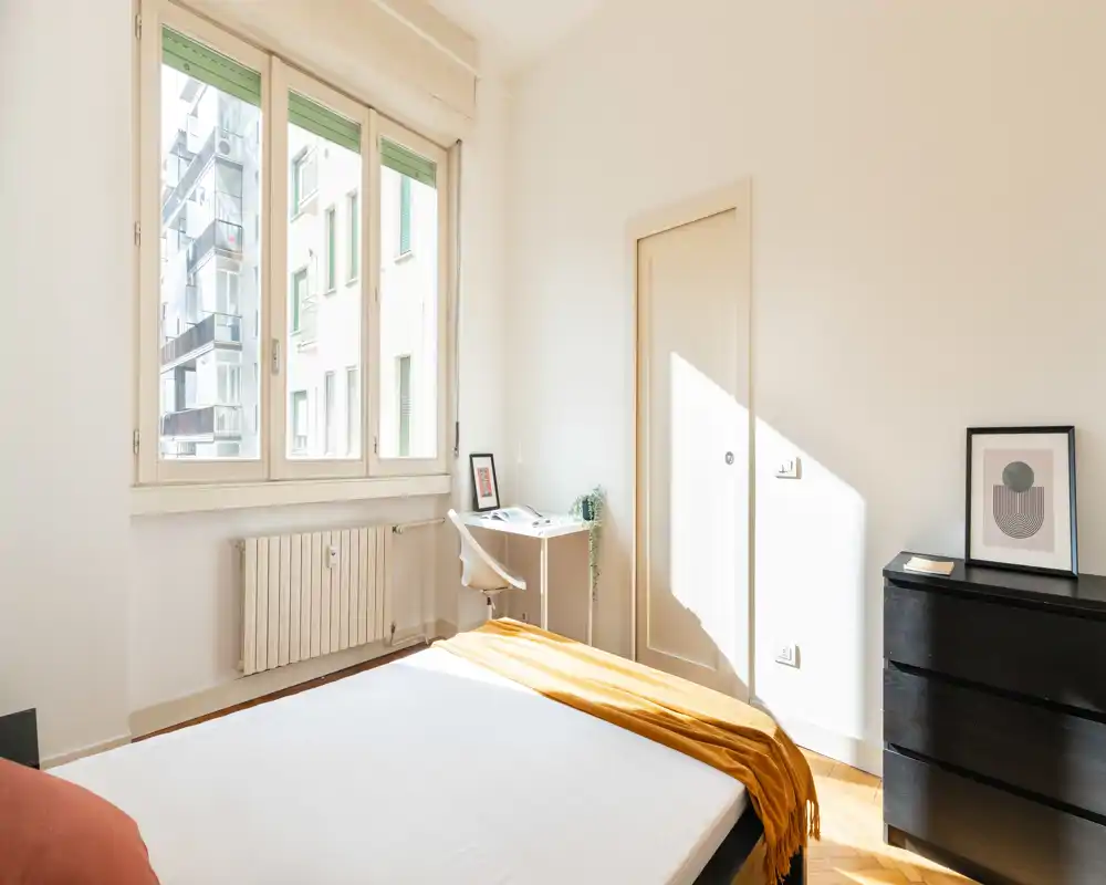 Dormitorio luminoso y moderno con cama bien hecha, ventana grande que aporta mucha luz natural, un pequeño escritorio junto a la ventana y una cómoda oscura. Paredes neutras y suelo de madera que crean un ambiente tranquilo y acogedor.