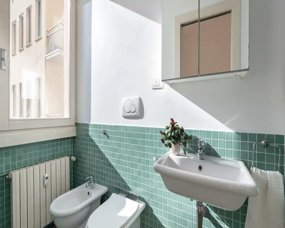 Baño moderno y luminoso con azulejos verdes tipo mosaico, lavabo mural, bidé y WC; amplia ventana que aporta luz natural y armario con espejo sobre el lavabo.