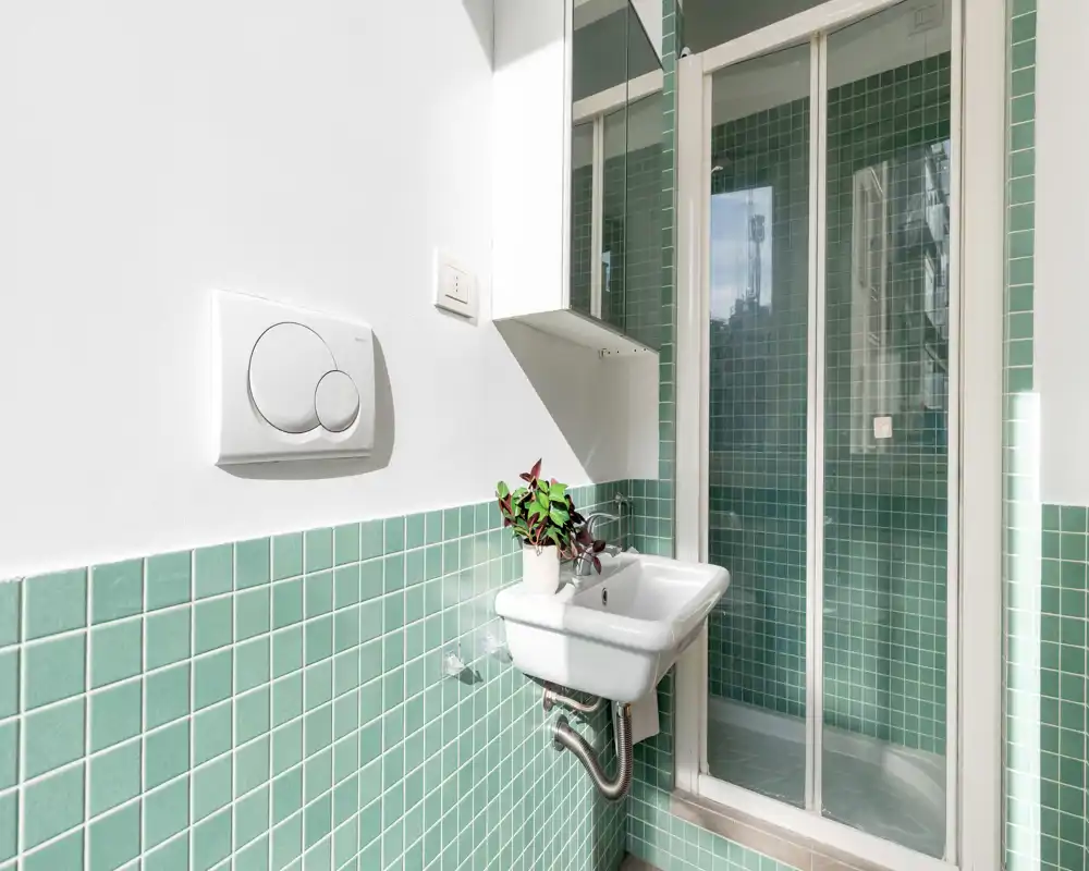 Rincón de baño luminoso con lavabo suspendido, azulejos cuadrados verdes y ducha con mampara de cristal.
