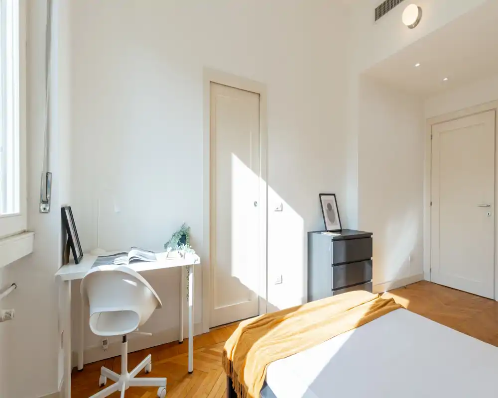 Dormitorio luminoso y minimalista con una cama acogedora con una manta cálida, un escritorio junto a la ventana y abundante luz natural.