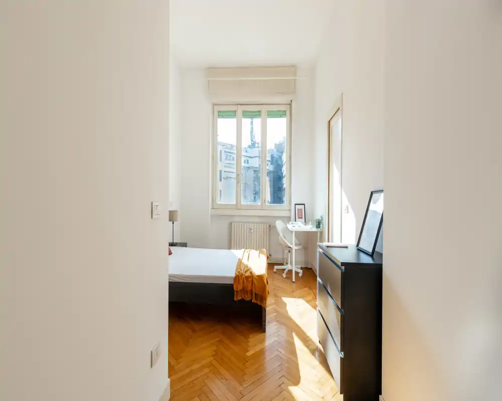 Dormitorio luminoso y minimalista con una gran ventana, suelo de parquet y cama bien hecha con una manta cálida al pie. Un pequeño escritorio junto a la ventana y una cómoda delgada completan el espacio aireado y funcional.