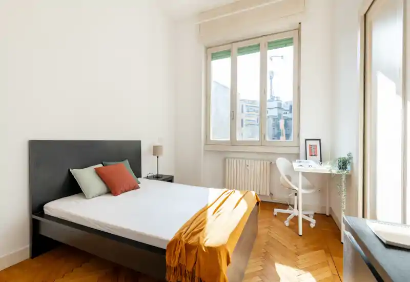 Dormitorio moderno y luminoso con cama doble, cojines decorativos y una manta cálida. Gran ventana que inunda la habitación de luz natural y un pequeño escritorio junto a la ventana como zona de trabajo.