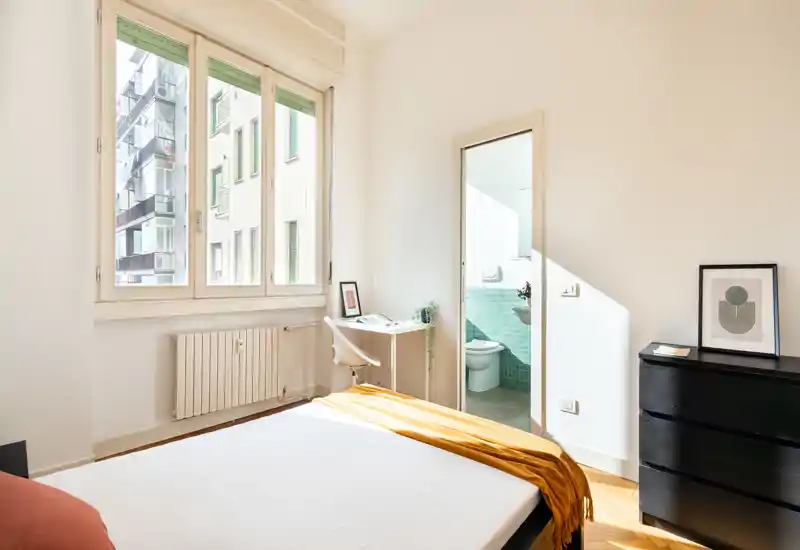 Dormitorio moderno y luminoso con una ventana grande, escritorio sencillo y baño privado. Estilo limpio, luz natural y textiles cálidos que crean un ambiente acogedor ideal para alquileres.