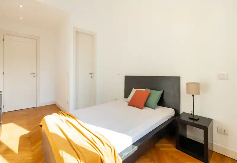 Dormitorio luminoso y minimalista con cabecero oscuro, cojines de colores y mesita con lámpara; el parquet cálido y la luz natural crean un ambiente acogedor.