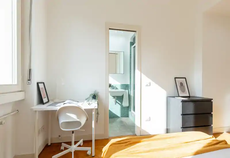 Dormitorio soleado con manta mostaza, un pequeño espacio de trabajo junto a la ventana y baño en suite con azulejos verdes — minimalista y acogedor.