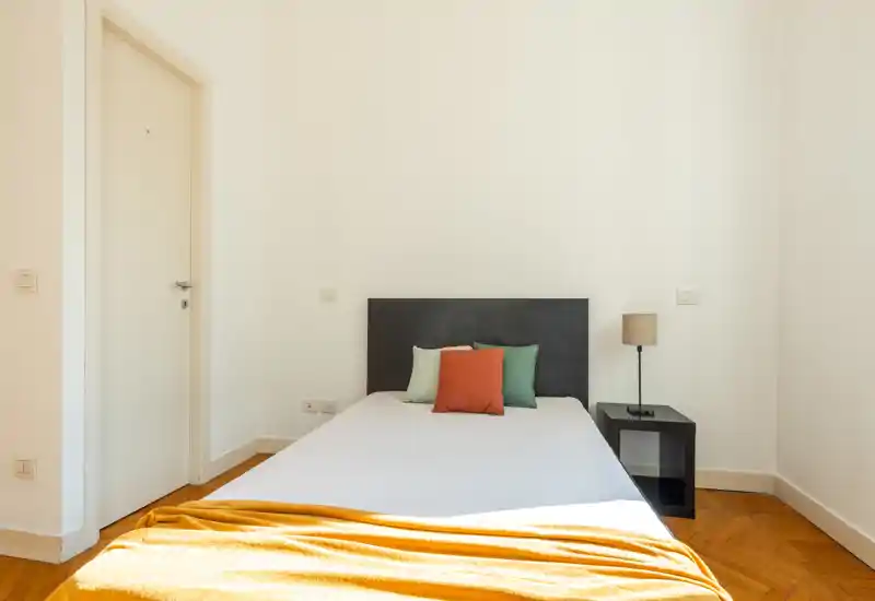 Dormitorio luminoso y minimalista con cama doble colocada en el centro, cabecero sencillo, mesita y lámpara; manta cálida y cojines decorativos que aportan calidez.