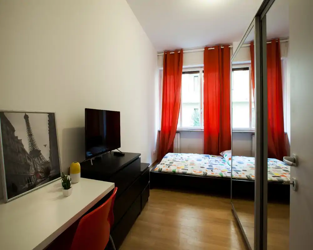 Camera da letto compatta e luminosa con biancheria a fantasia, tende rosse calde e armadio con ante a specchio che amplia visivamente lo spazio. Piccola scrivania e TV per soggiorni pratici.