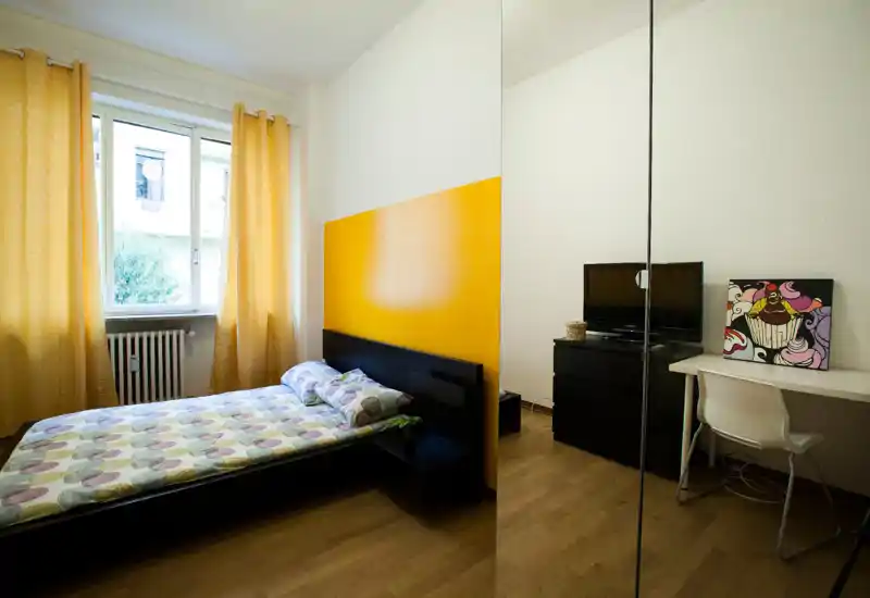 Camera da letto luminosa e accogliente con letto matrimoniale davanti a una parete gialla, ampia finestra con tende calde e armadio con specchio che amplia lo spazio. Scrivania e comò con TV sul lato opposto la rendono funzionale.