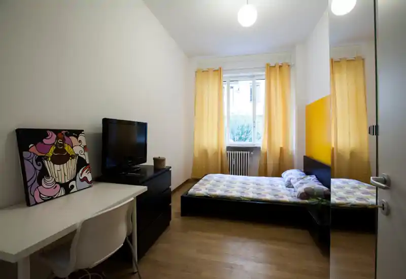 Camera da letto luminosa e ordinata con letto basso matrimoniale, biancheria con motivo e tende gialle calde. Un armadio con porta specchiata e una scrivania con TV rendono lo spazio funzionale e adatto a brevi soggiorni.