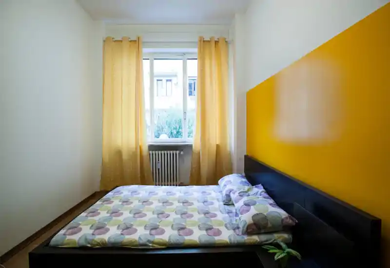 Camera da letto luminosa e ordinata con letto matrimoniale, biancheria a motivi e una parete gialla come elemento caldo; la luce naturale dalla finestra rende l'ambiente accogliente.