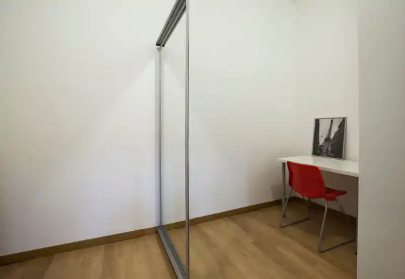 Petit coin de pièce minimaliste avec un bureau blanc, une chaise rouge et une armoire coulissante miroir ; lumineux et prêt à être personnalisé.