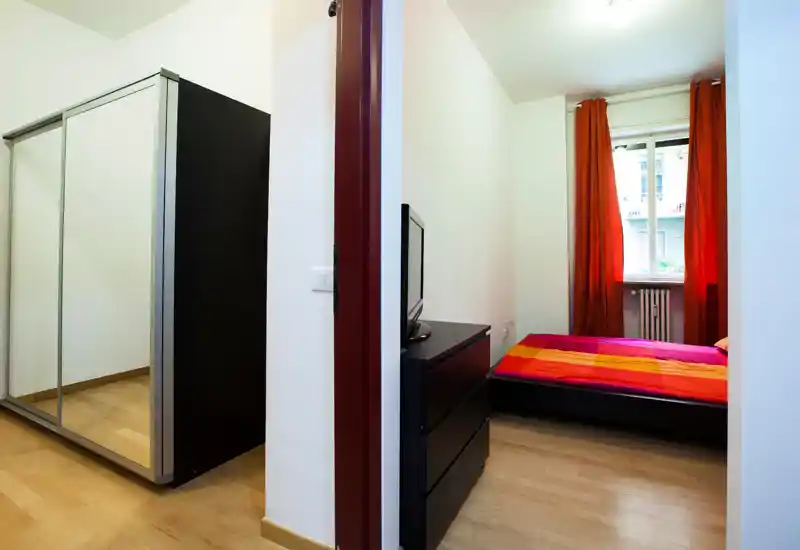 Chambre compacte et lumineuse avec lit bas habillé de linge rouge-orangé, commode avec TV et armoire à miroirs offrant un rangement pratique.