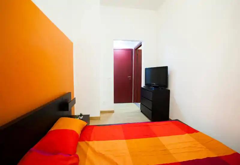 Chambre lumineuse et moderne avec un grand lit aux draps orange et rouge, une tête de lit sombre et une commode avec télévision face au lit. La décoration épurée et les couleurs chaudes rendent l'espace convivial.
