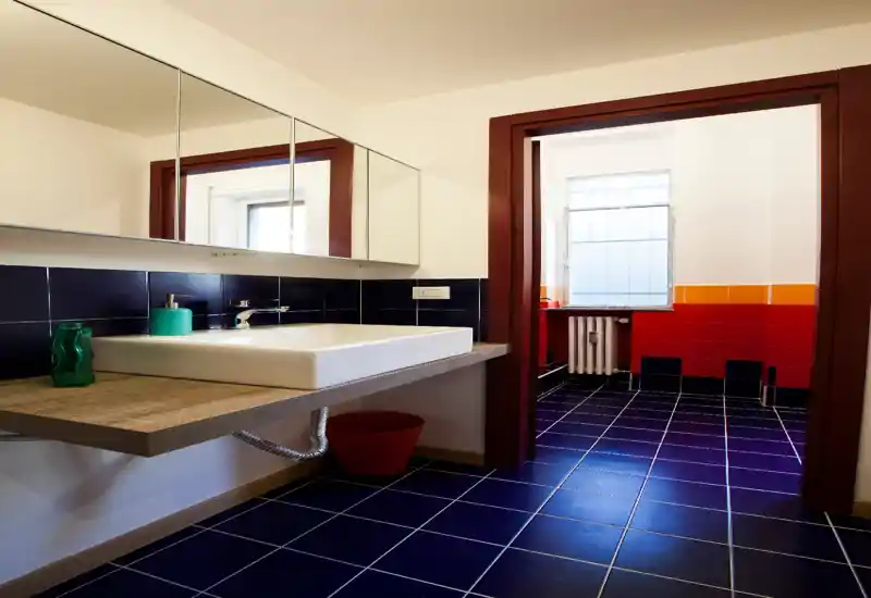 Bright, modern bathroom with a rectangular countertop sink, wall-mounted mirror cabinet and deep blue floor tiles contrasted by red and orange wall tiles in an adjacent section.