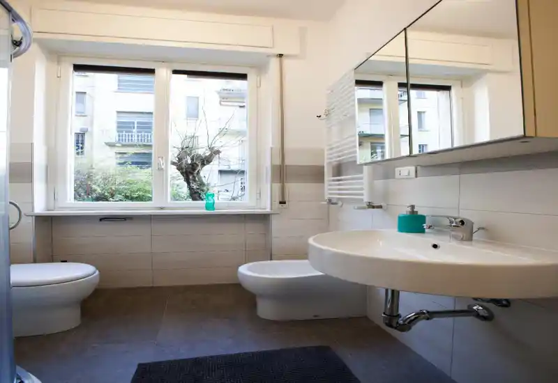 Bright, modern bathroom with a wall-mounted sink, bidet and toilet, a large window providing natural light, and a mirrored cabinet above the sink.