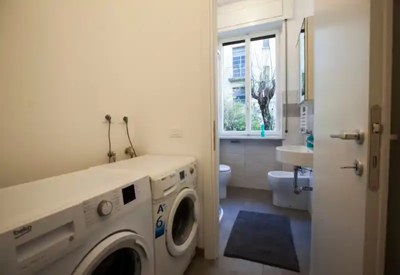 Compact bathroom with sink, bidet and toilet, bright natural light from a large window and a tidy mat on the floor. A laundry area with washer and dryer sits just outside the doorway.