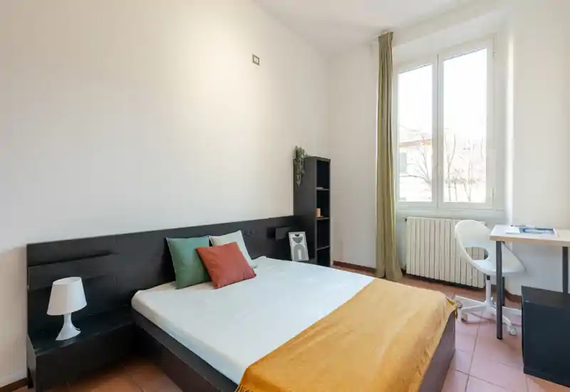 Dormitorio luminoso y minimalista con cama doble, cojines de colores y plaid mostaza; gran ventana que aporta mucha luz natural y un pequeño escritorio junto al radiador.