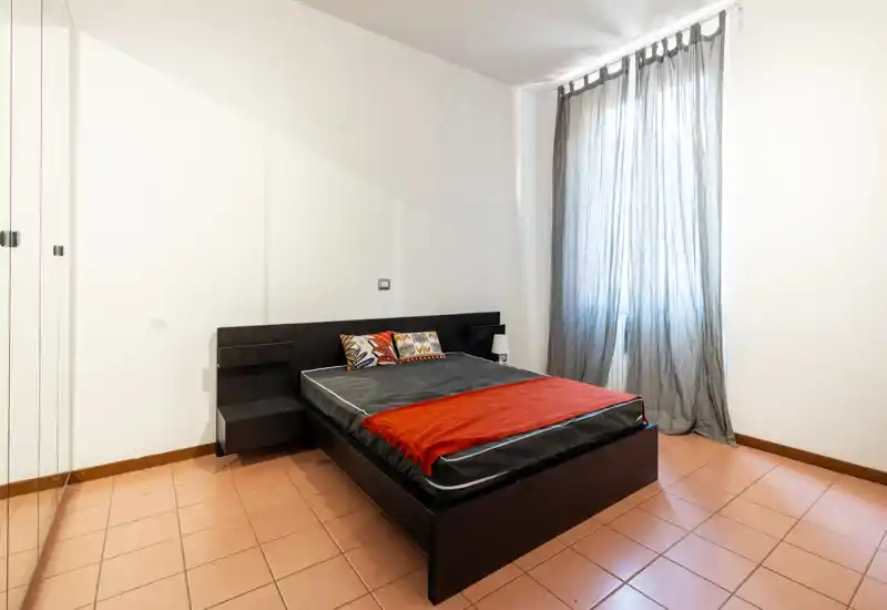 Chambre lumineuse et minimaliste avec un lit double à cadre sombre, coussins décoratifs et jeté rouge; grande fenêtre avec voilage laissant entrer la lumière naturelle.