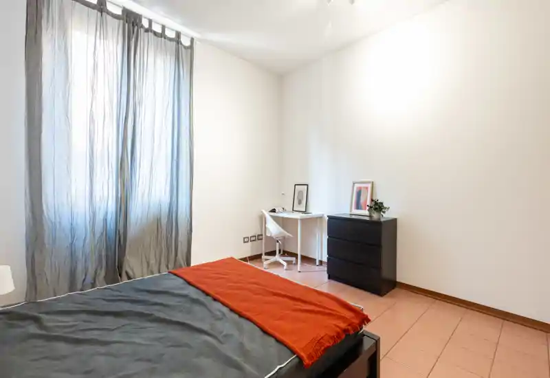 Chambre lumineuse et minimaliste avec un lit double en linge foncé et un plaid couleur rouille. La lumière naturelle traverse des rideaux légers; un petit bureau et une commode noire complètent l'aménagement.