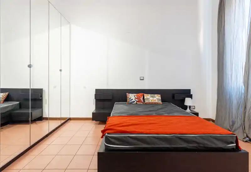 Chambre minimaliste avec lit bas en bois foncé, coussins colorés et plaid orange; la penderie miroir reflète la pièce et agrandit l'espace perçu. La lumière naturelle apporte une ambiance moderne et nette, idéale pour une annonce locative.