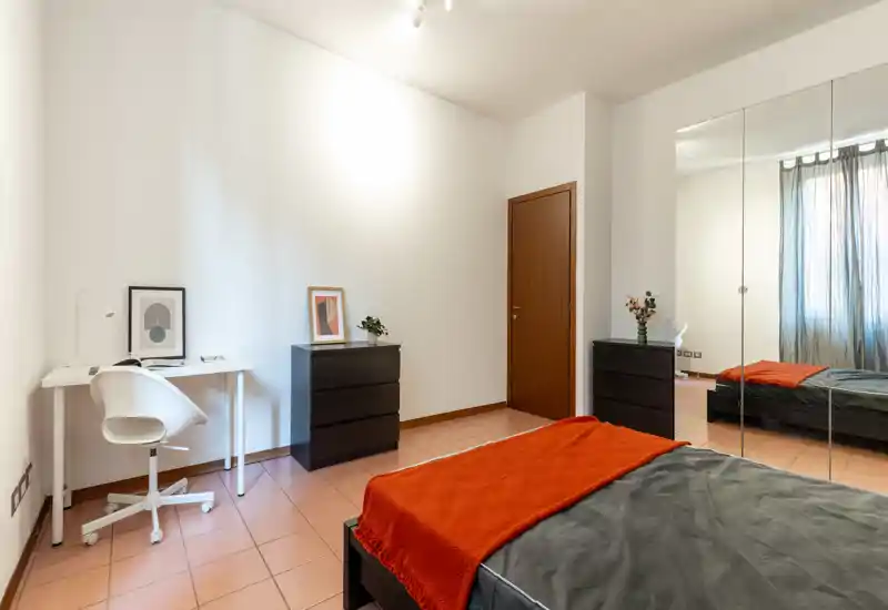 Chambre moderne et lumineuse avec lit en linge sombre et plaid orange, grande armoire à portes miroir qui apporte de la profondeur et un petit bureau — idéale pour une personne ou un étudiant.