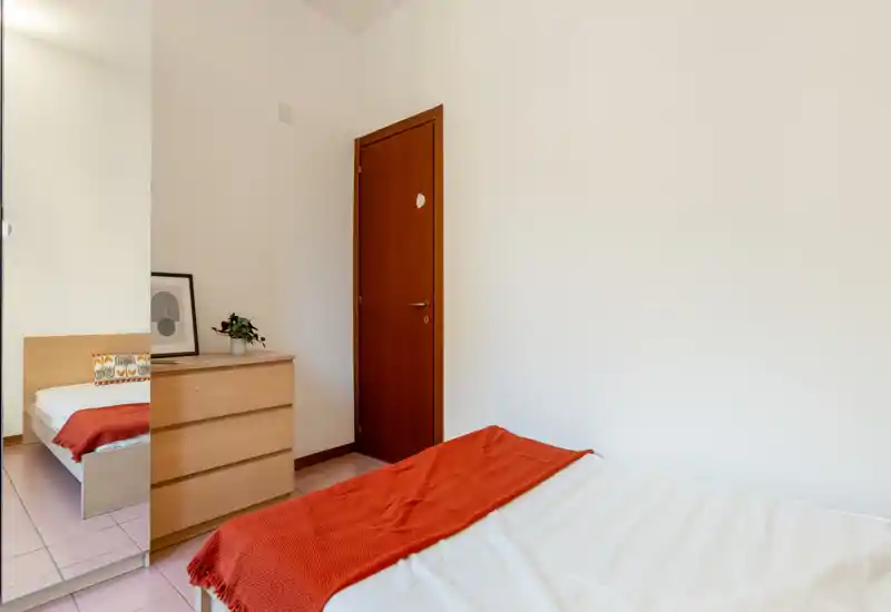 Chambre minimaliste et lumineuse avec lit double en linge blanc, plaid couleur rouille, commode en bois et miroir pleine longueur reflétant une partie du lit.