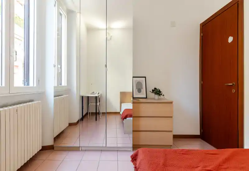 Chambre lumineuse et minimaliste avec grandes fenêtres, armoire miroir, commode en bois et lit partiellement visible avec une couverture rouge chaleureuse.