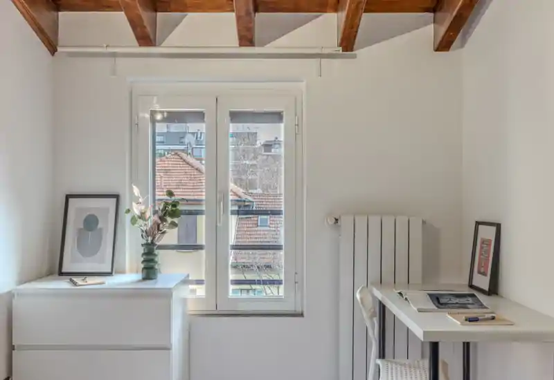 A bright, minimal corner with a window, small desk and dresser — styled with simple decor and wooden ceiling beams. The photo highlights a workspace and storage area rather than a clear bedroom or living room.