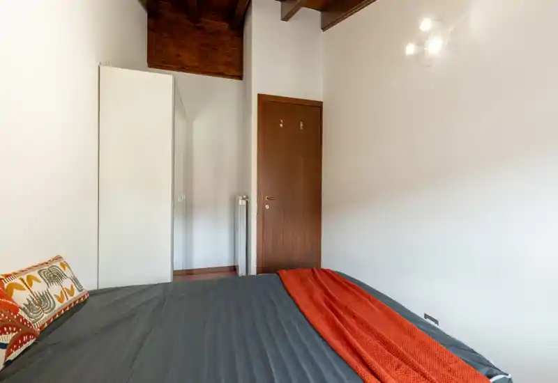 Bright, minimal bedroom with exposed wooden ceiling beams, a large bed dressed in dark linens and a red throw, plus a white wardrobe and wooden door.