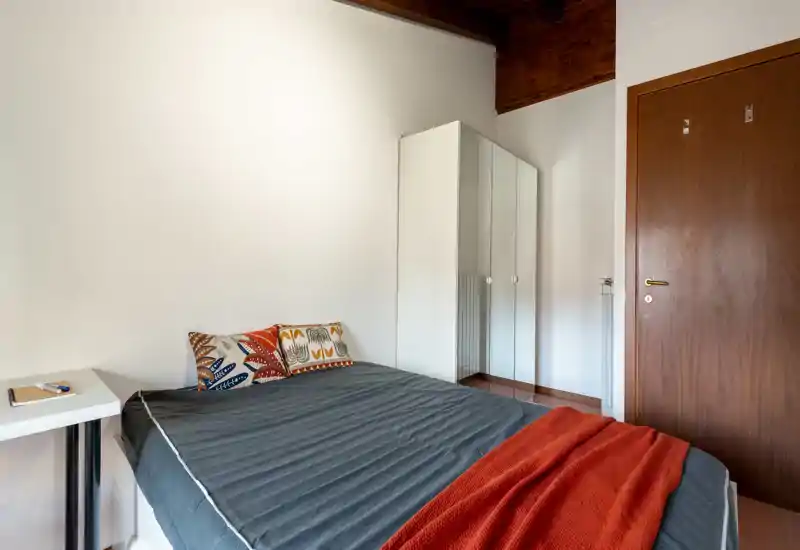 Bright, simply styled bedroom with a double bed dressed in dark linens, colorful decorative pillows and a warm orange throw; mirrored wardrobe and wooden ceiling beam add character.