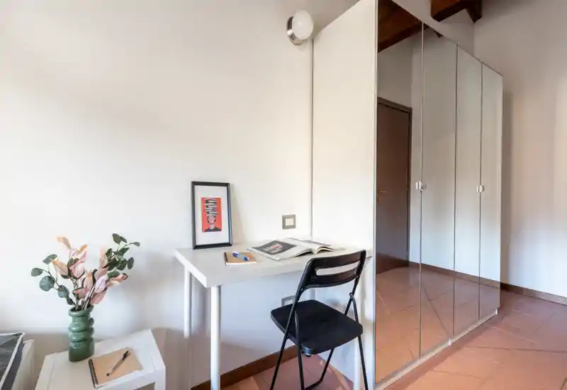 Angolo d'ingresso compatto con una piccola scrivania, sedia pieghevole e guardaroba con ante a specchio che riflettono il corridoio. Spazio pulito e luminoso adatto come area d'accoglienza o postazione veloce.