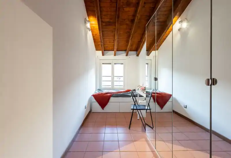 Camera da letto mansardata con soffitto in legno inclinato, letto su piattaforma vicino alla finestra, coperta rossa e cuscini, e armadio con ante a specchio che ampliano lo spazio.