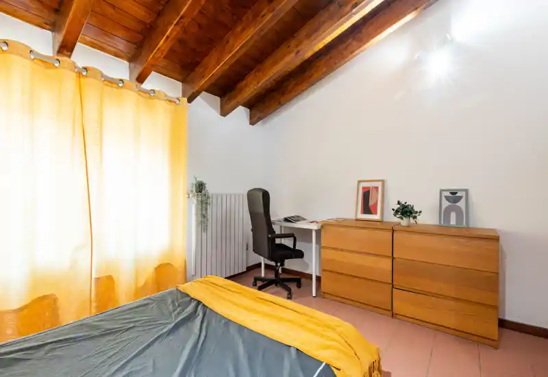Bright, cozy bedroom with exposed wooden beams, a neatly made bed in the foreground and a small desk with dresser along the wall — warm textiles and natural light create a welcoming atmosphere.
