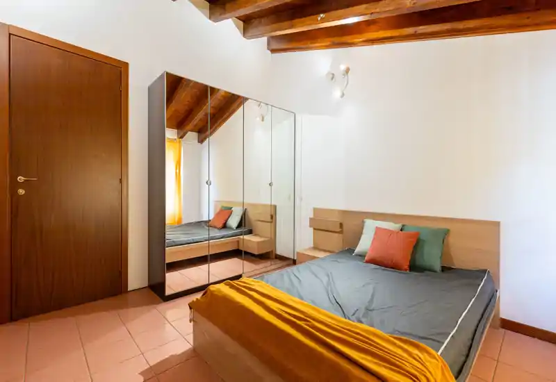Bright, modern bedroom with a double bed, colorful cushions and warm wooden ceiling beams. A mirrored wardrobe and bedside tables complete the space, ideal for a comfortable stay.