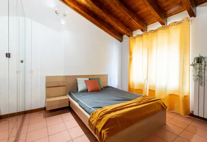 Cozy bedroom with a wooden slatted ceiling, double bed with cushions and a warm mustard throw, and soft natural light through yellow curtains.