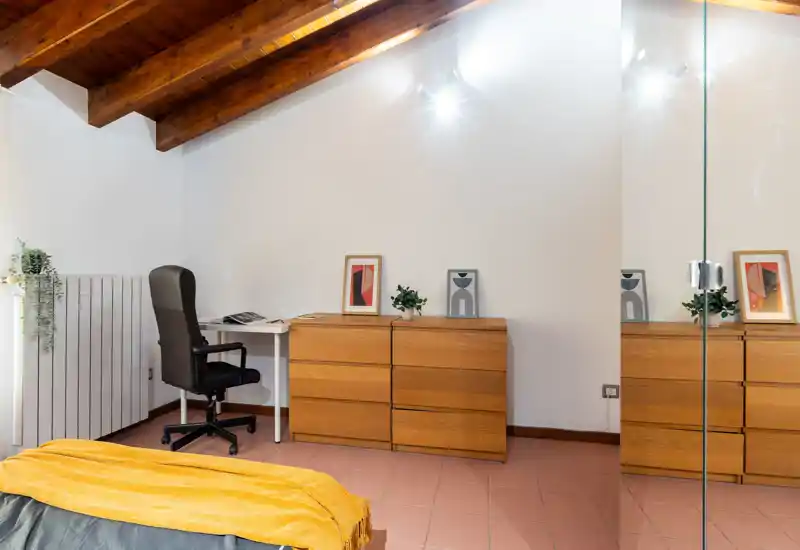 Coin chambre mansardée avec la couverture du lit partiellement visible au premier plan, poutres en bois, commode, bureau et chaise — lumineux, bien stylé et adapté aux annonces locatives.