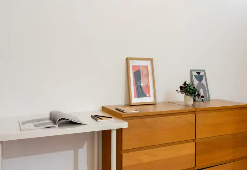 Coin de chambre minimaliste avec une commode en bois, des cadres et une petite plante sur le dessus ; décor lumineux, propre et moderne, idéal pour des photos de mise en valeur.