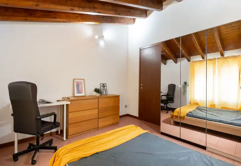 Bright, cozy bedroom with exposed wooden beams, a double bed dressed in a grey duvet and yellow throw, mirrored wardrobe and a small desk — ideal for restful stays and remote work.