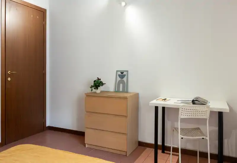Rincón de dormitorio sencillo y funcional con cómoda de madera, escritorio y vista parcial de la cama con una manta cálida. Decoración limpia y minimalista con luz suave, apta para un anuncio de alquiler.