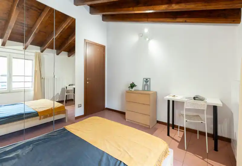 Bright, neatly styled bedroom with exposed wooden beams, a double bed with warm throw and a desk by the wall — ideal for a comfortable stay or a work-from-home setup.