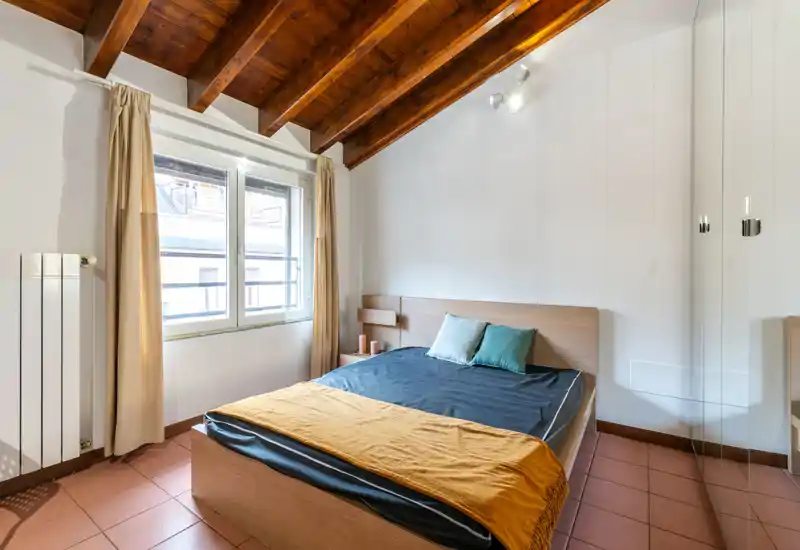 Bright, cozy bedroom with a wooden slatted ceiling, double bed, two decorative cushions and a mustard throw. Large window provides natural light and the simple modern headboard and tiled floor give a clean, welcoming look ideal for rental listings.