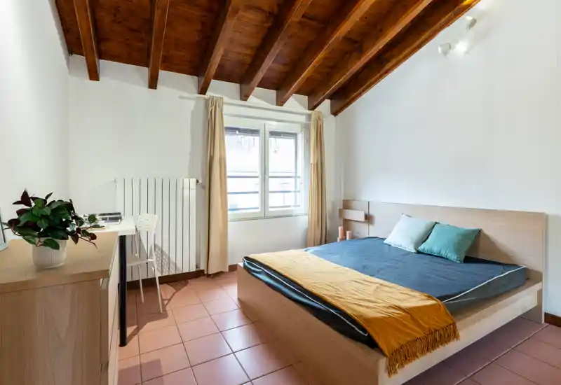Bright, well-composed bedroom with a wooden-beamed ceiling, a double bed dressed with a dark duvet and a mustard throw, plus a small desk and dresser by the window.