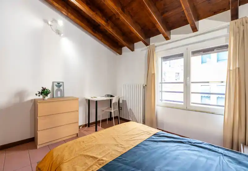 Bright, cozy bedroom with wooden vaulted ceiling, a double bed with mustard and blue linens, a small study desk and chest of drawers next to a large window.