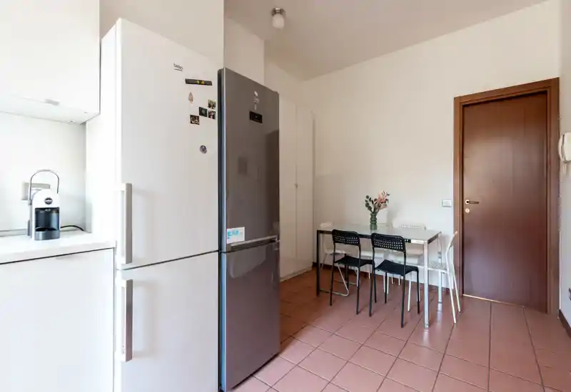 Cocina compacta y ordenada con frigoríficos dobles, cafetera y una pequeña mesa para cuatro. Tonos neutros y luz natural que crean un espacio funcional y agradable.