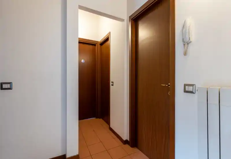 Entrada/pasillo corto de apartamento con suelo de terracota, puertas de madera y portero automático en la pared — espacio de tránsito funcional.